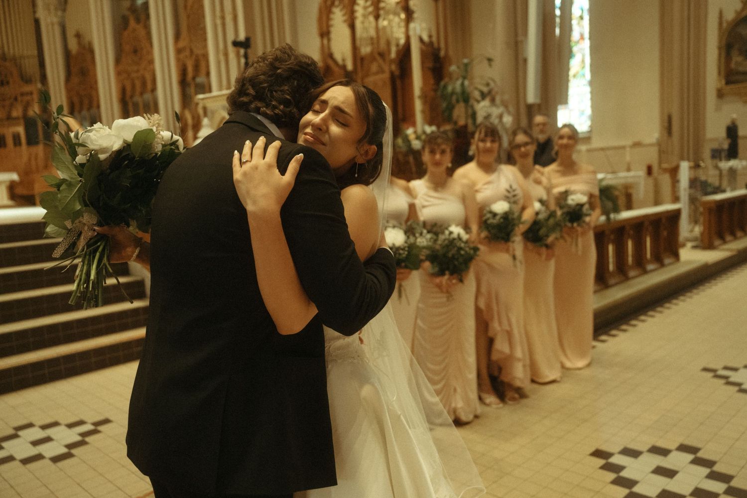 Bride in white gown embracing guest while bridesmaids in champagne dresses hold bouquets in church.