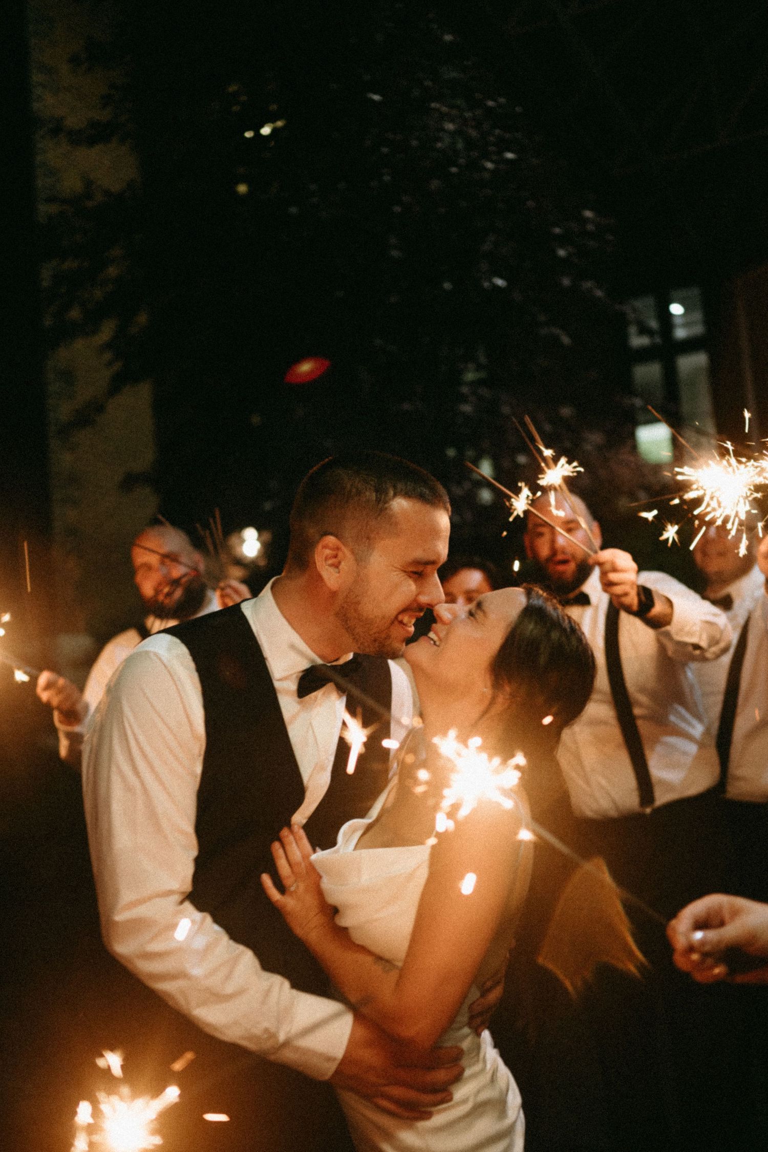 Bride and groom share a romantic moment surrounded by guests holding sparklers at a night wedding celebration.