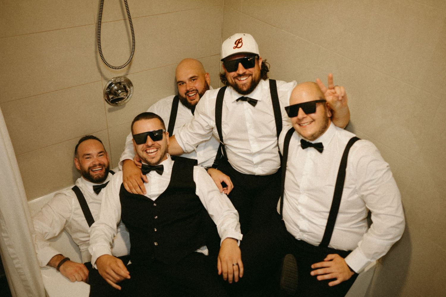 Five groomsmen in white shirts, bow ties, and suspenders pose together in a bathroom, smiling and giving thumbs up.