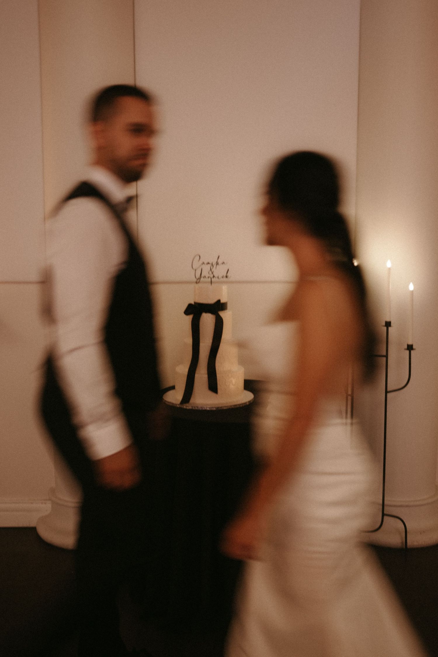 Blurred bride and groom walking past a white wedding cake with black bow at elegant indoor reception.