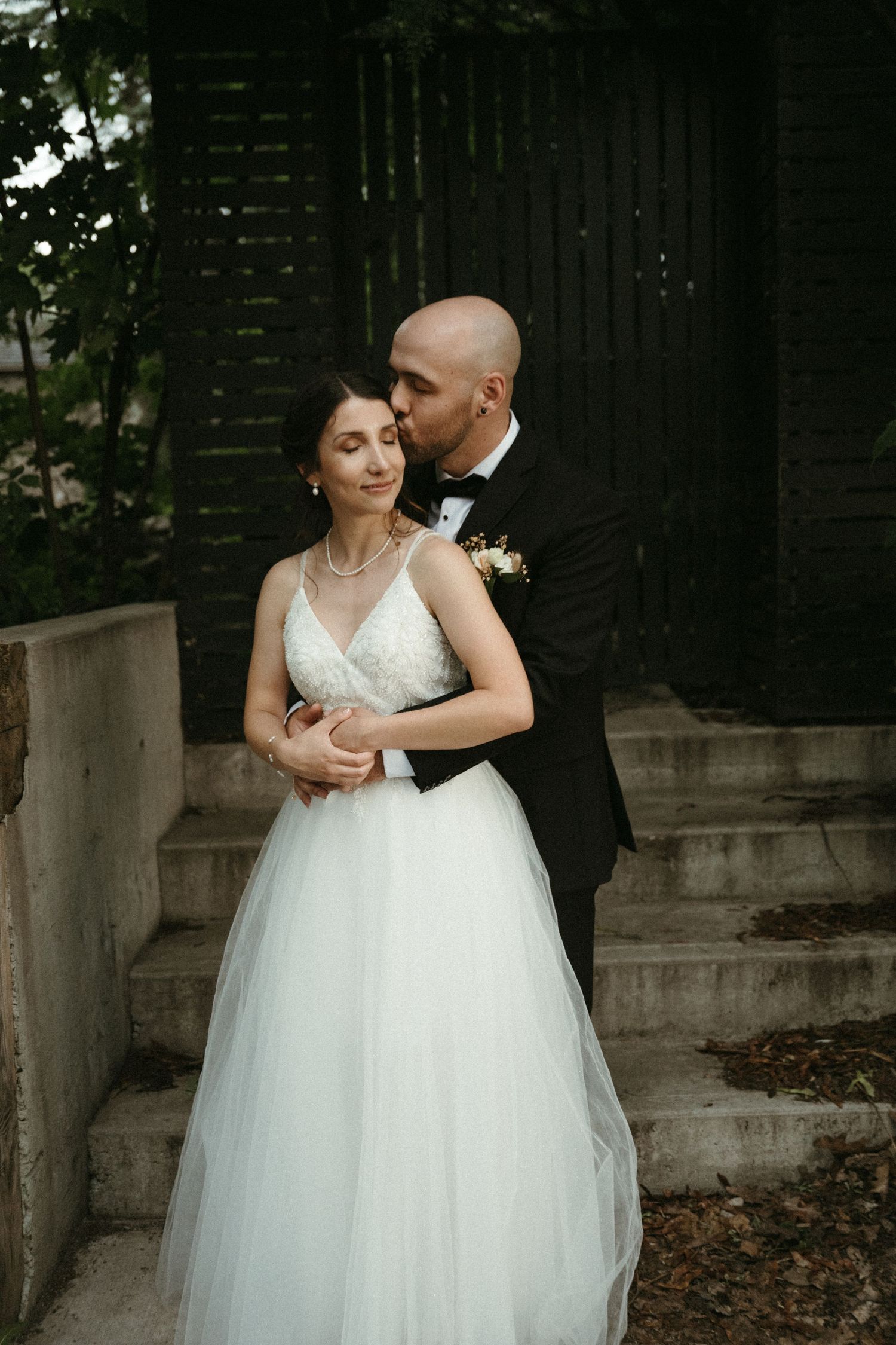 Groom in black tuxedo tenderly kissing bride in lace tulle gown on stone steps.