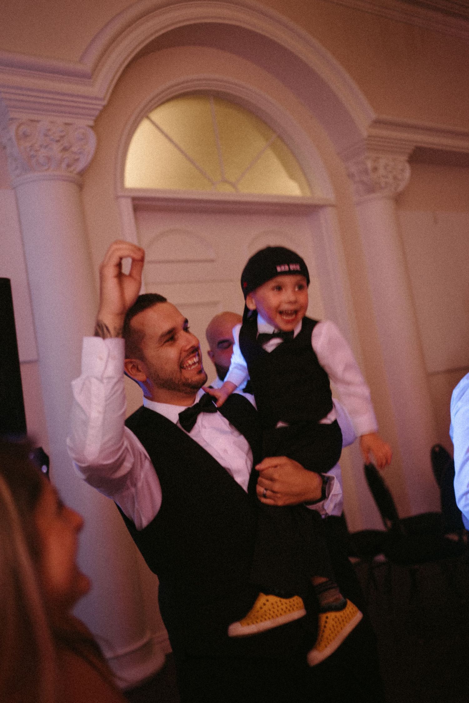 Man in tuxedo joyfully lifts smiling boy in vest and cap at elegant wedding reception.