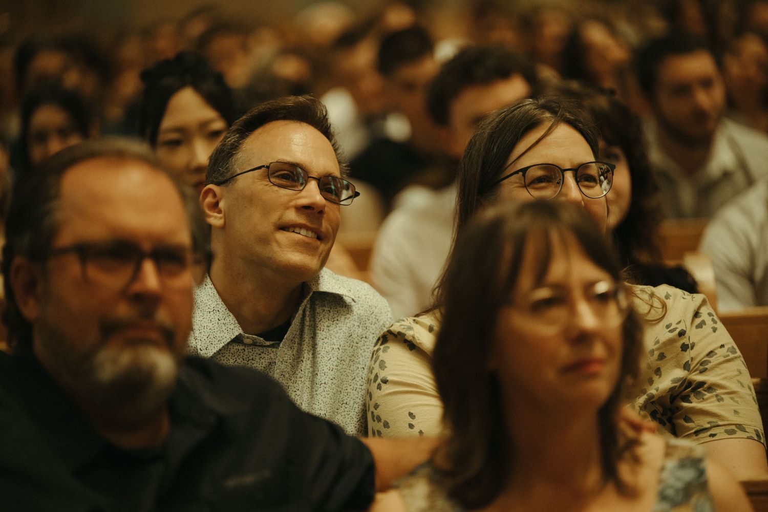 Smiling wedding guests wearing glasses seated in a crowded church during a wedding ceremony.