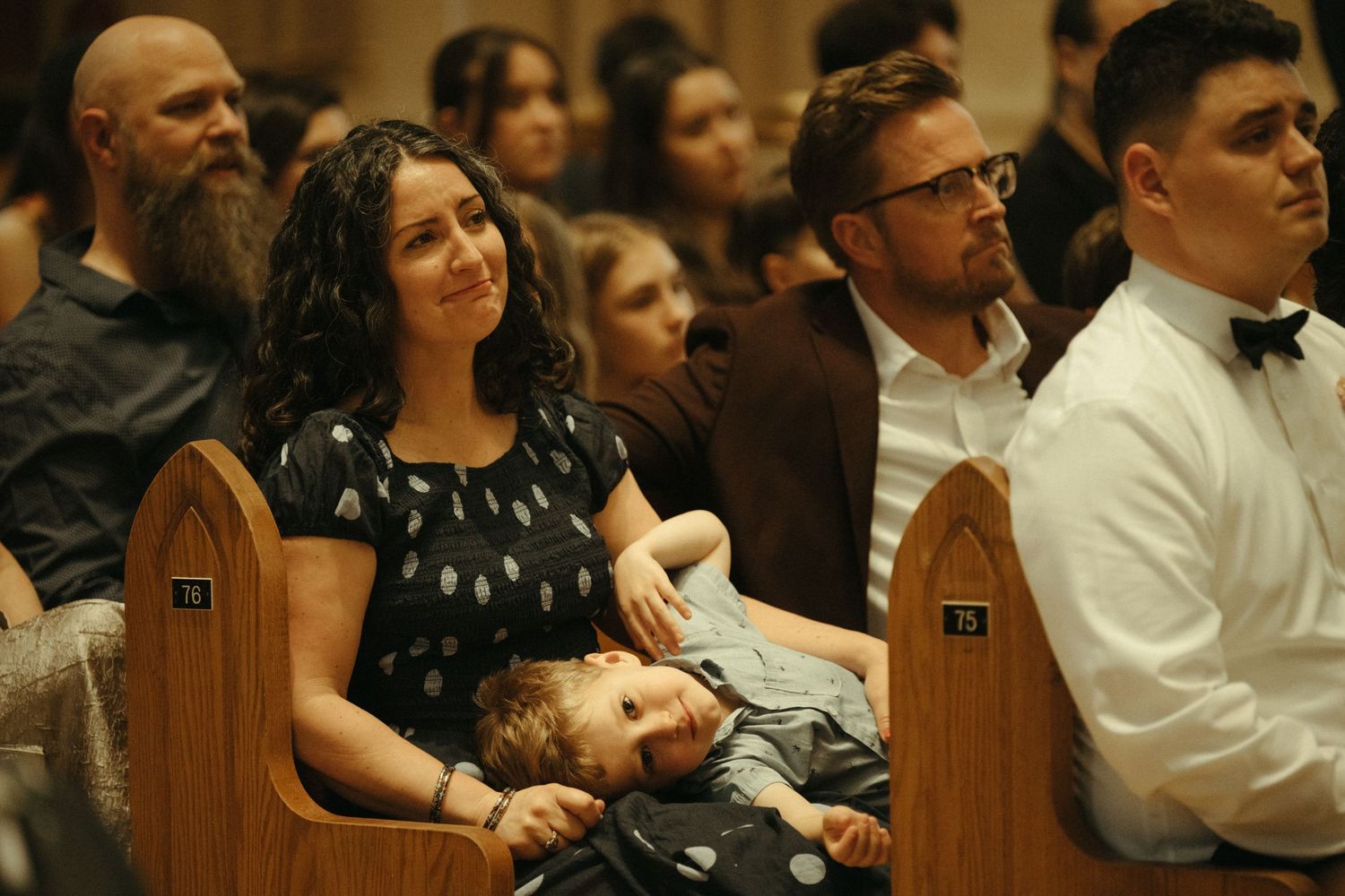 Wedding guest cradling a sleeping toddler in church pew while watching the ceremony with a smile.