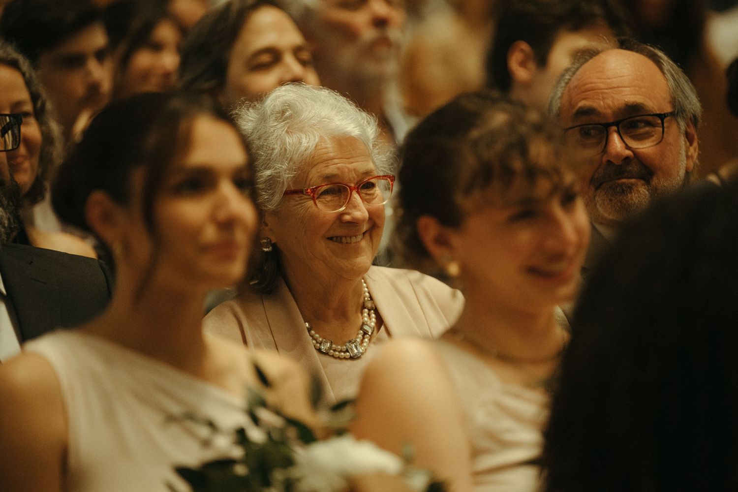 Smiling elderly woman with pearl necklace enjoys wedding ceremony surrounded by guests.