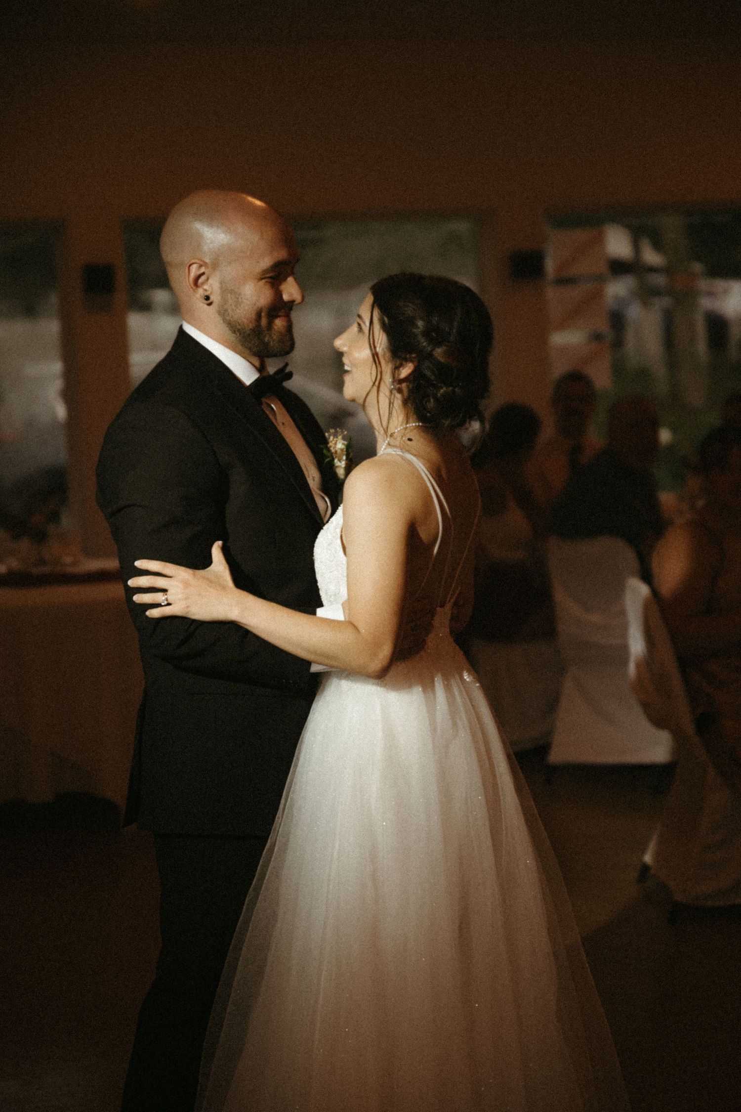 Bride and groom share romantic first dance in dimly lit reception hall, gazing lovingly at each other.