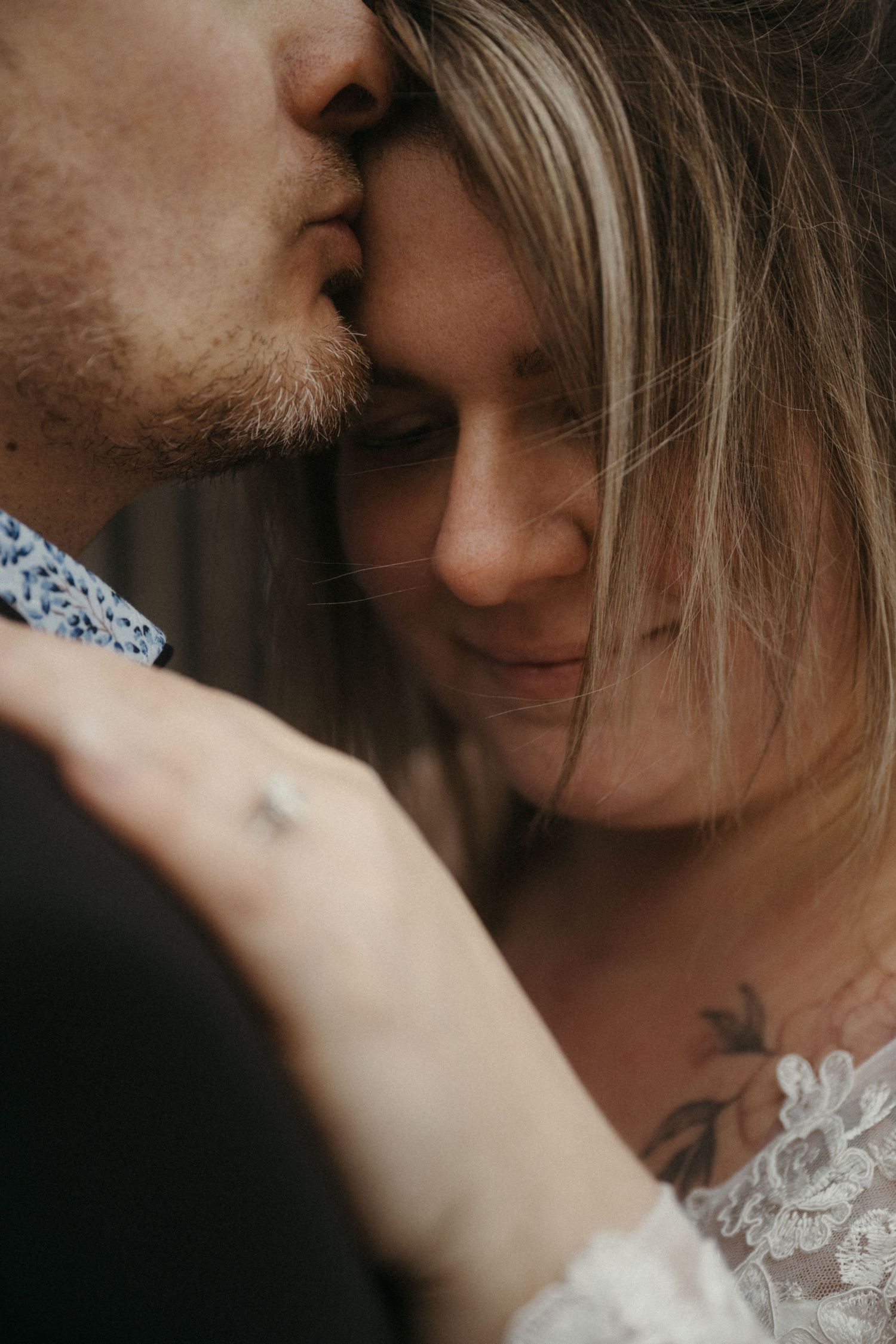 Groom kisses bride on forehead in a close romantic portrait showing her floral chest tattoo.