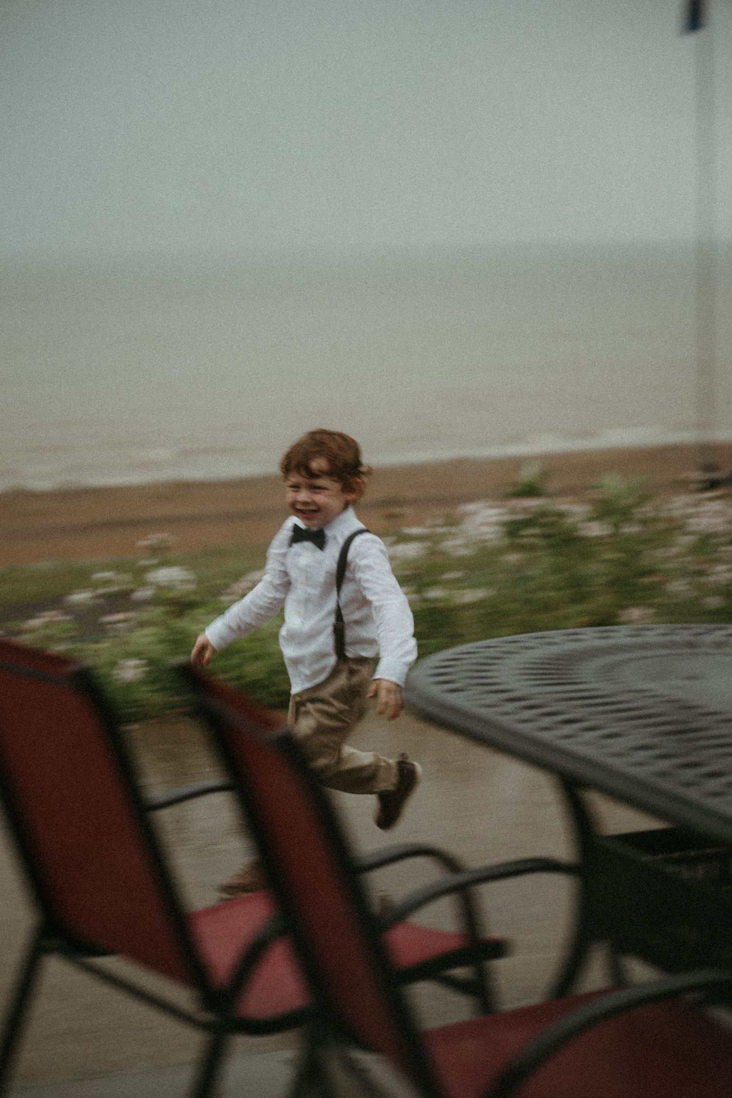 Joyful young ring bearer in white shirt and suspenders running near seaside patio at wedding.
