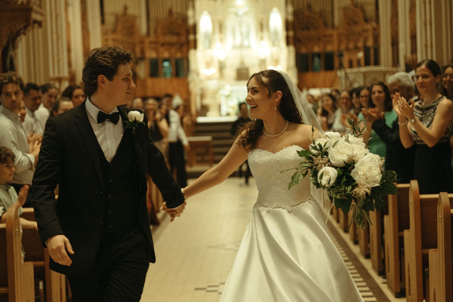 Newlyweds walk down church aisle smiling, bride holding white bouquet, guests applauding in background.