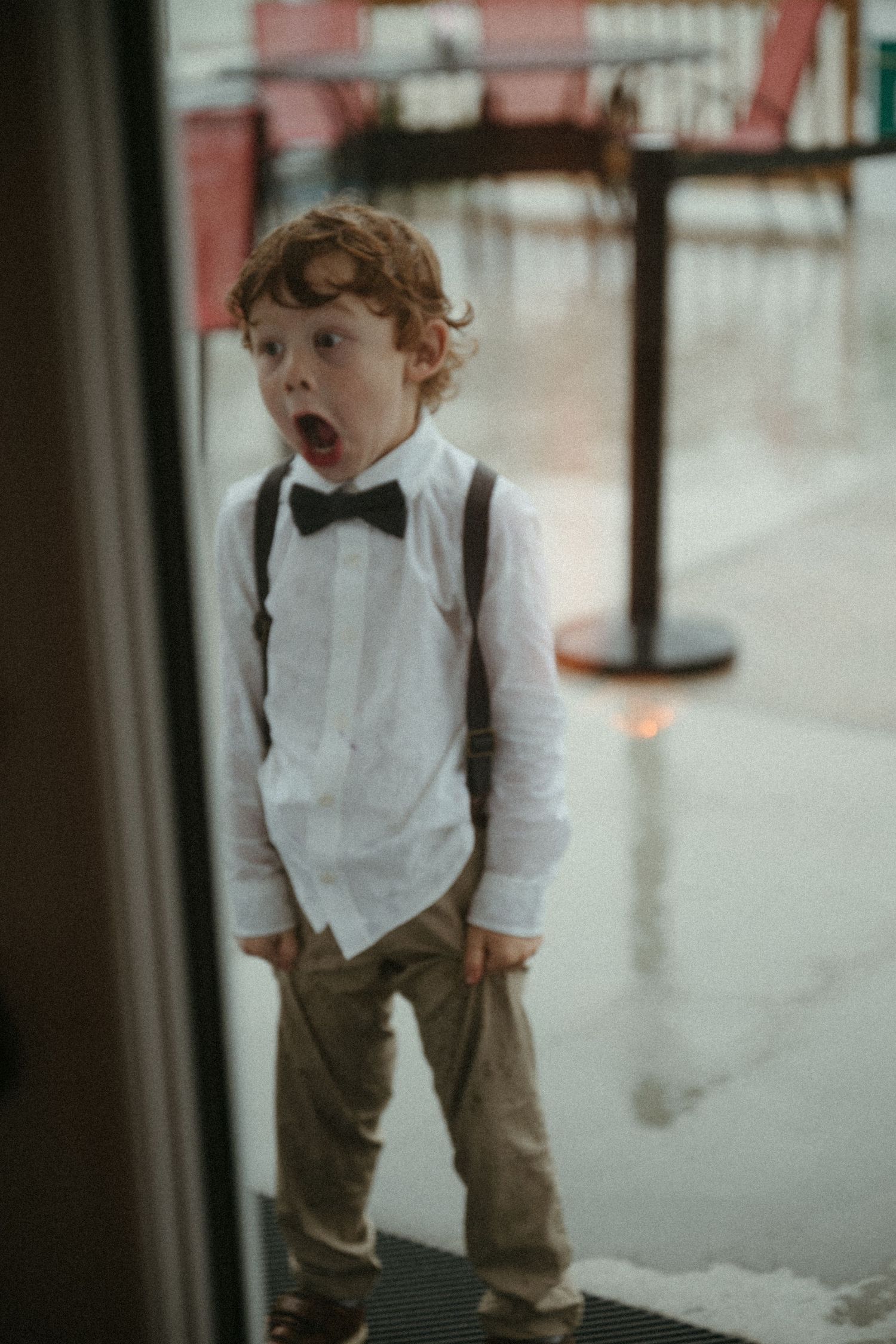 Adorable redhead ring bearer boy in bow tie suspenders and khakis with surprised expression.