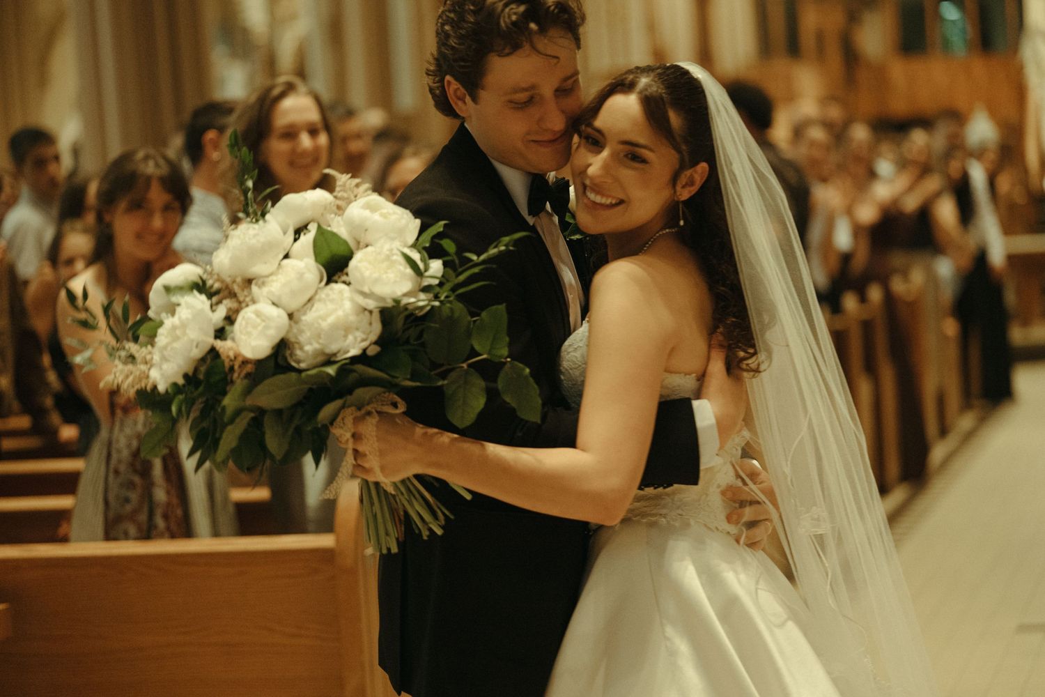 Joyful newlyweds embrace in church aisle, bride holding white rose bouquet after ceremony.