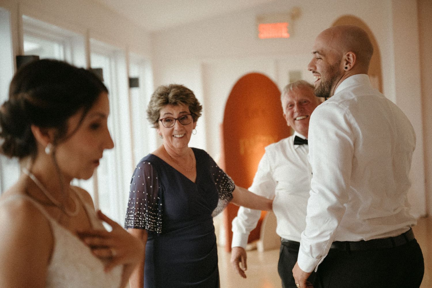 Bald groom laughs with elderly woman in navy dress while bride stands nearby indoors.