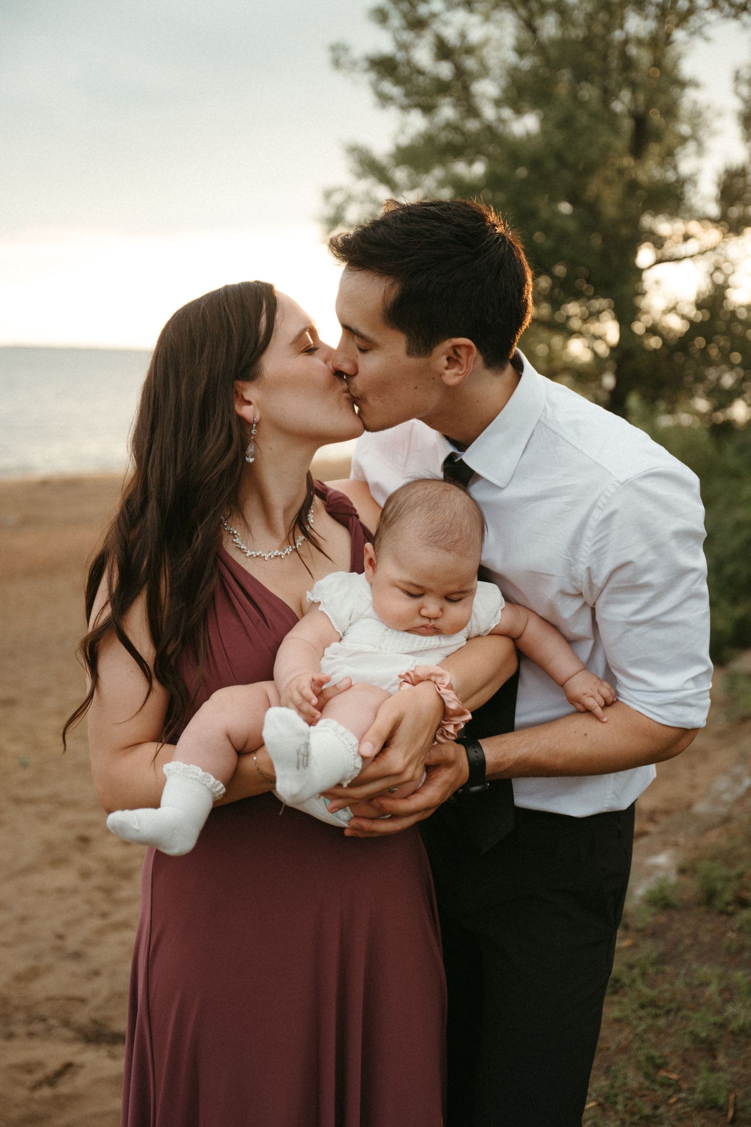 A couple kisses on a beach at sunset while holding their newborn baby dressed in white.