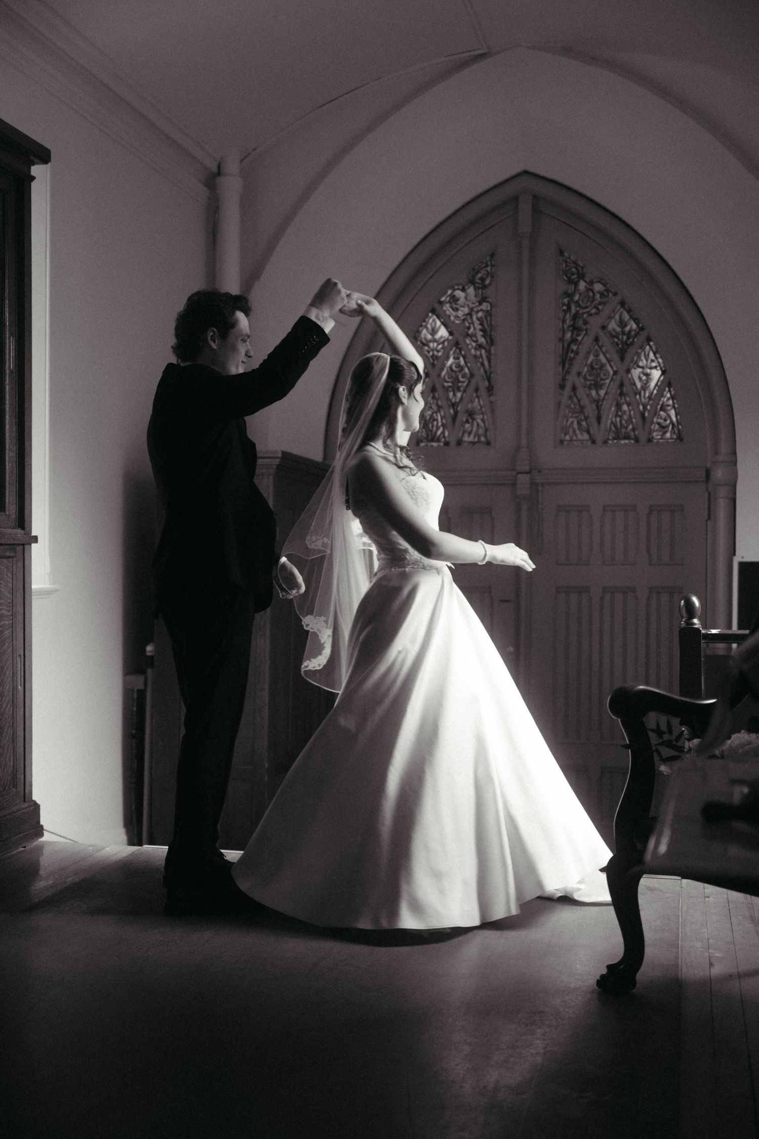 Black and white photo of a groom twirling his bride in a wedding dress inside a chapel with stained glass windows.