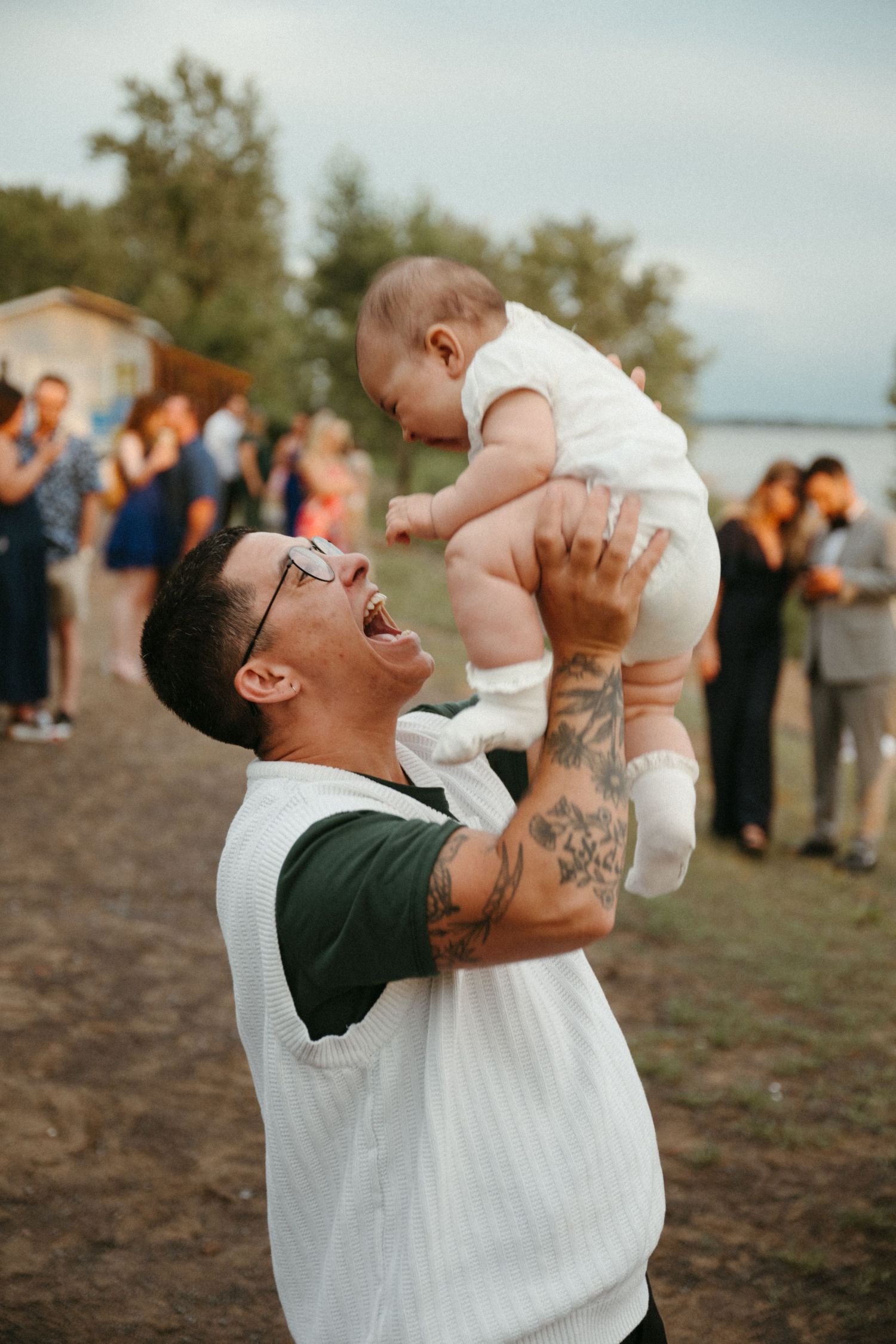 Tattooed man with glasses joyfully lifting a baby in the air at an outdoor wedding reception.