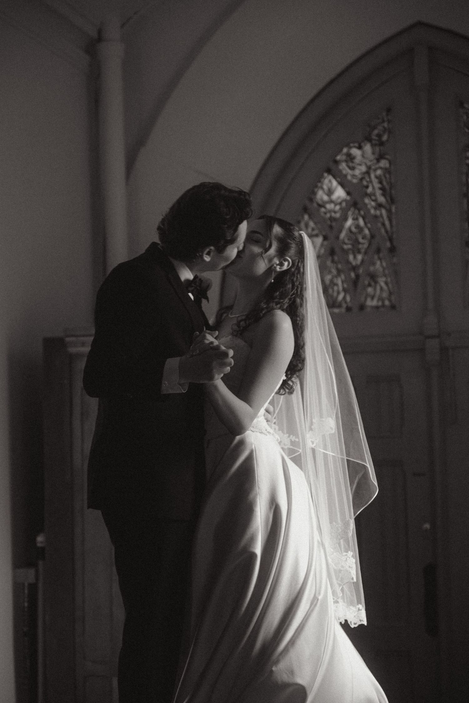Black and white photo of a bride and groom sharing a first kiss in a church, with stained glass windows in the background.