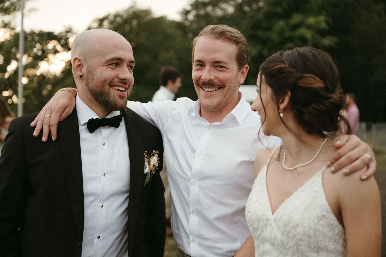Groom in tuxedo and bride in white wedding dress smile with a guest outdoors at a wedding reception.