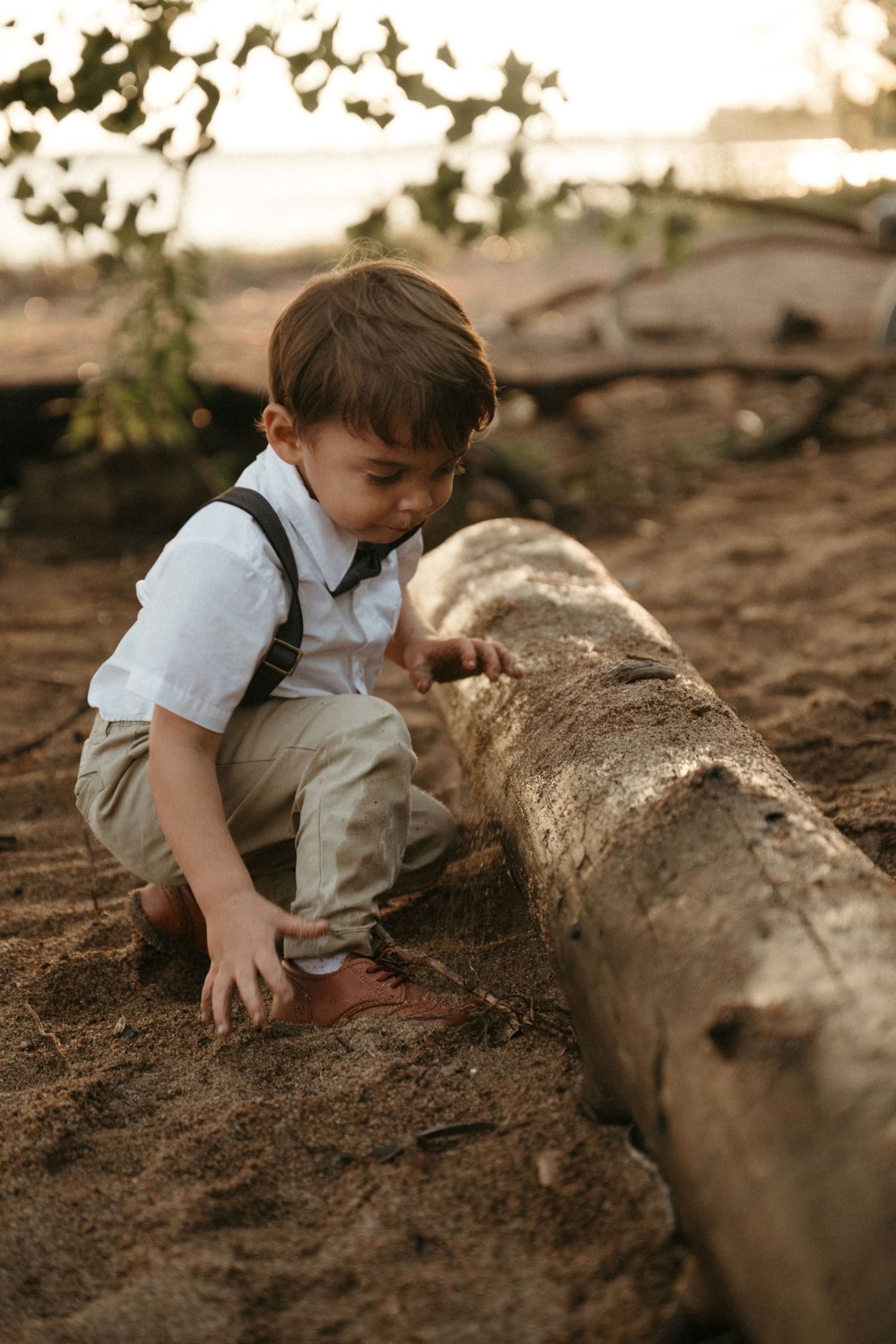 A young boy in suspenders crouches barefoot on sandy ground, curiously exploring a weathered log outdoors.