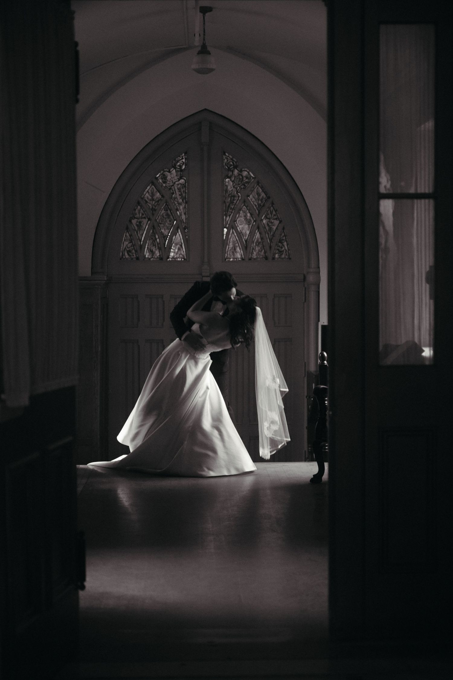 Bride and groom sharing a romantic kiss in a dimly lit church with stunning stained glass windows in black and white.