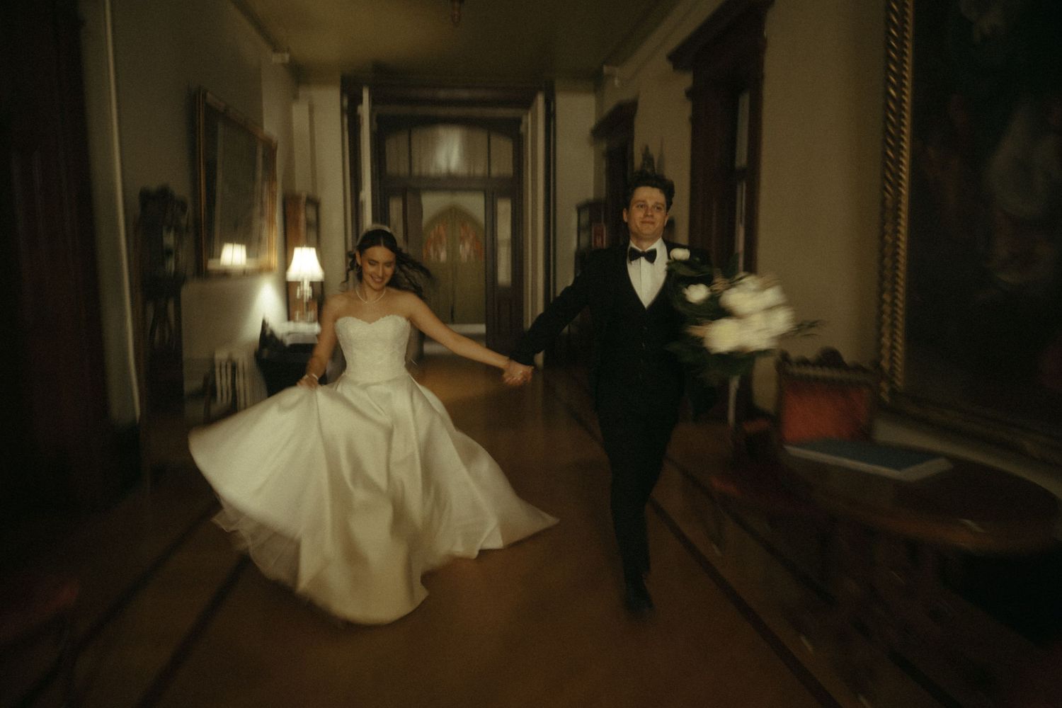 A joyful bride and groom run hand-in-hand through an ornate hallway, bride holding a white bouquet.