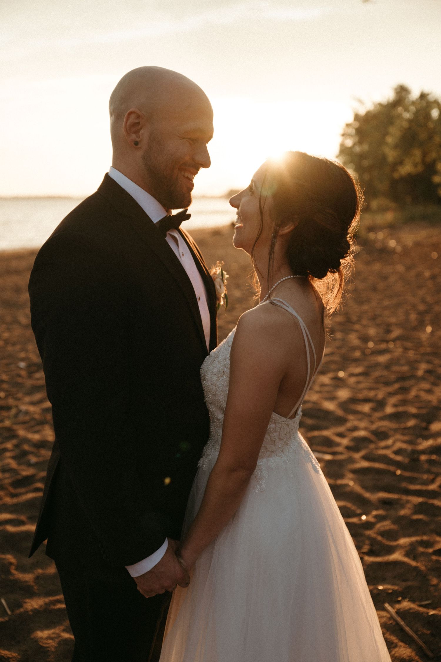 A bride and groom hold hands on a sandy beach at golden sunset, smiling at each other in wedding attire.