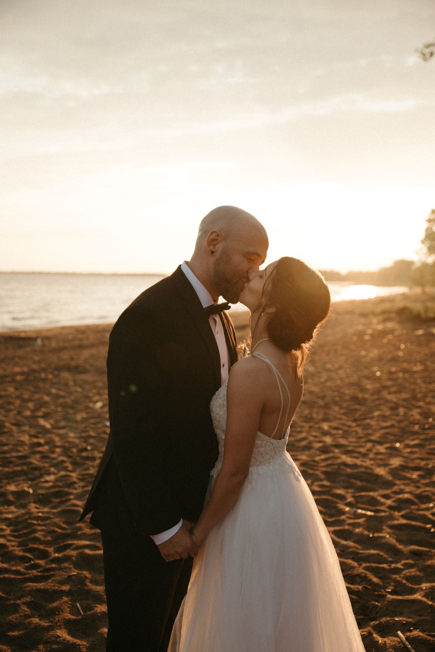 A bride and groom share a romantic kiss on a sandy beach at golden sunset, with the ocean glowing behind them.