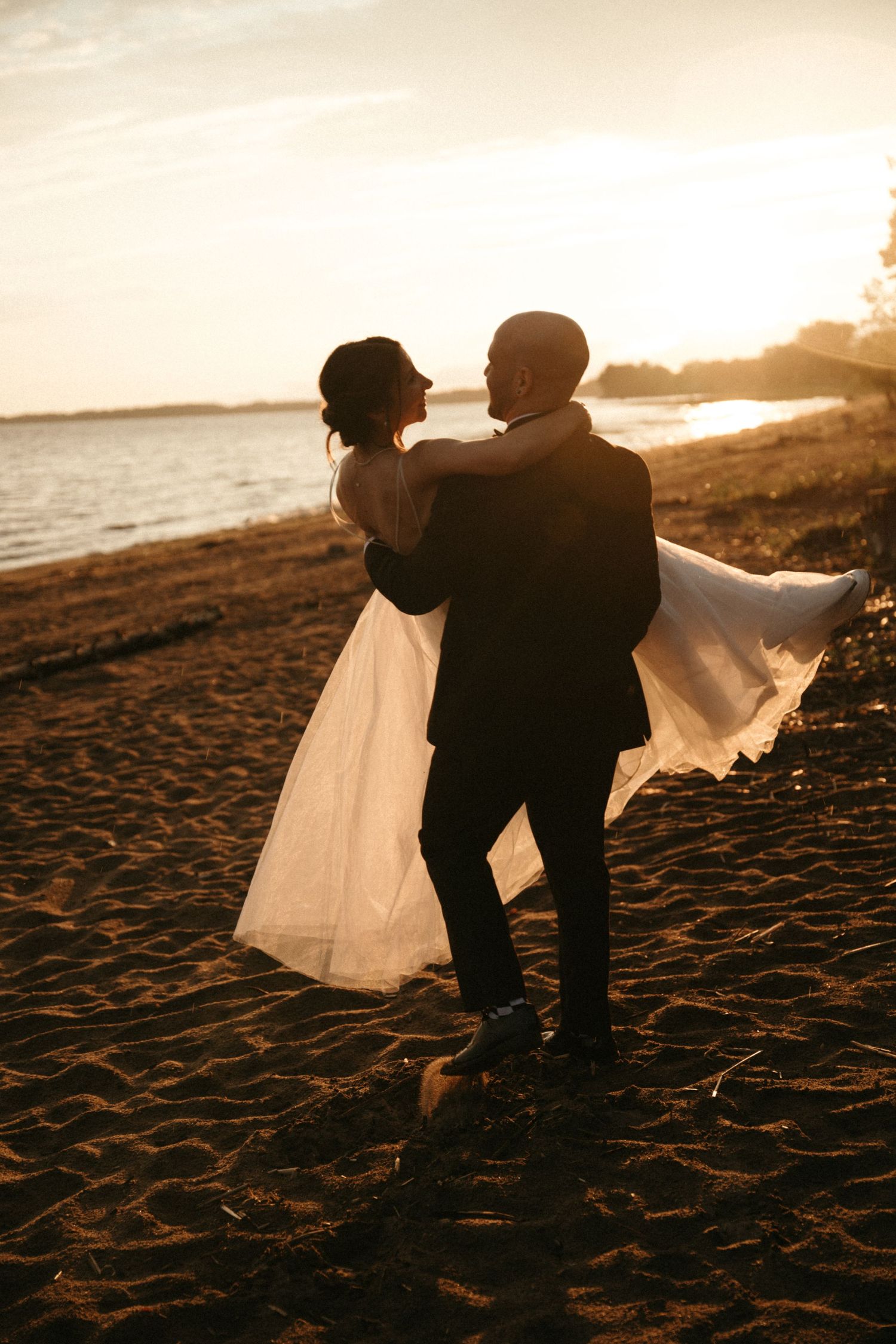 A groom lifts his bride in a flowing white dress on a sandy beach at golden sunset.