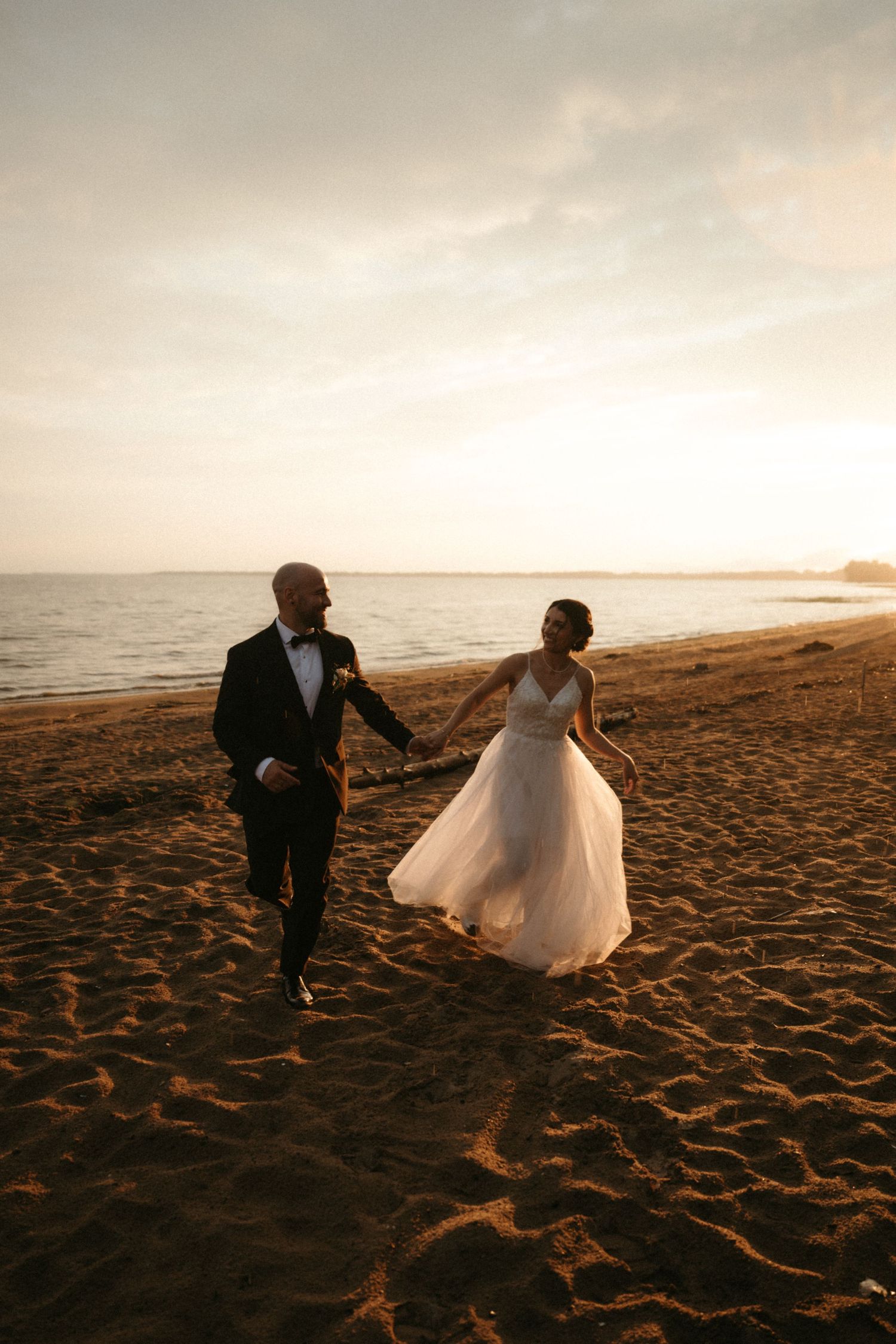 Bride and groom holding hands running on a sandy beach at golden sunset during their wedding day.