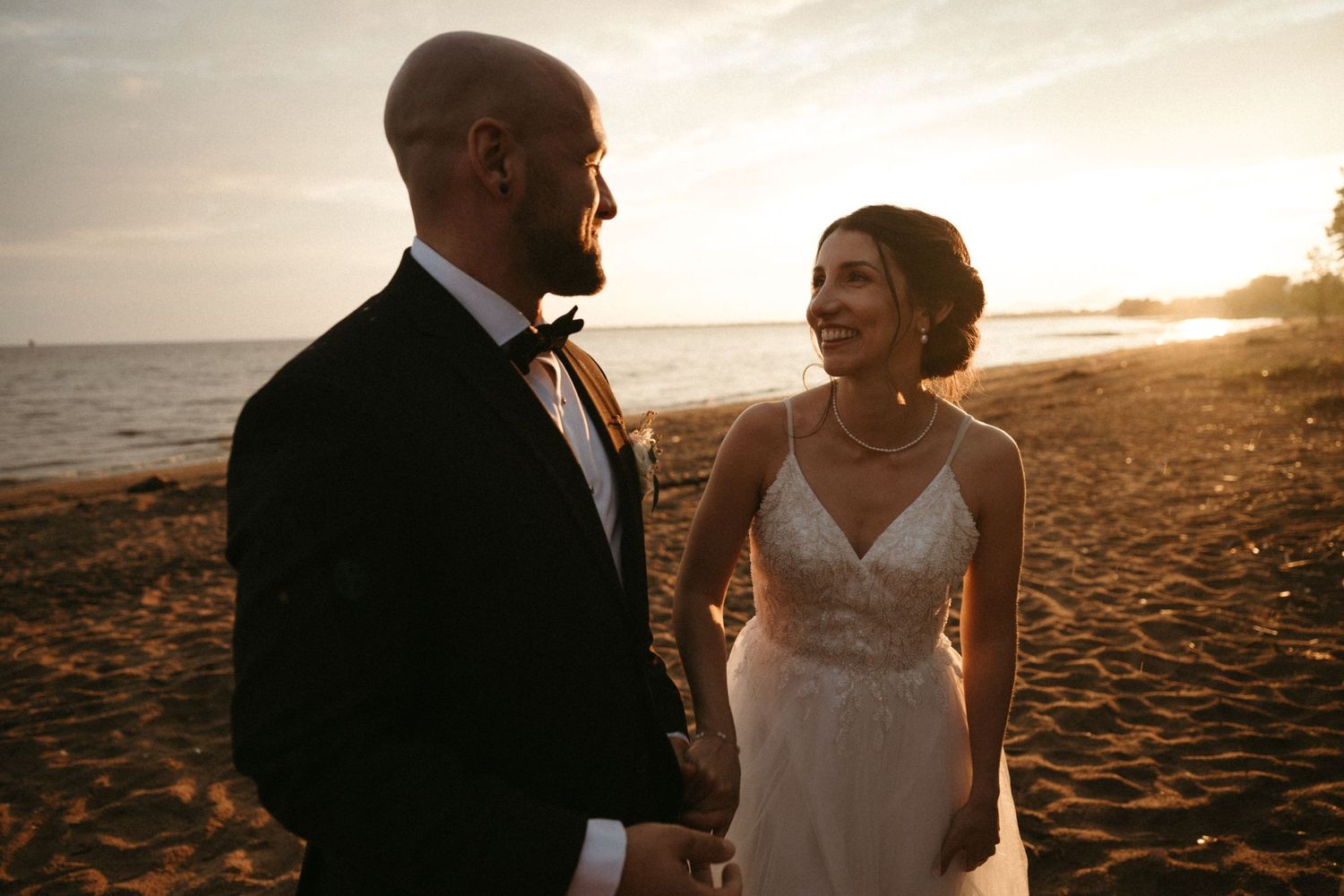 Bride and groom holding hands on a sandy beach at golden sunset, smiling at each other in wedding attire.