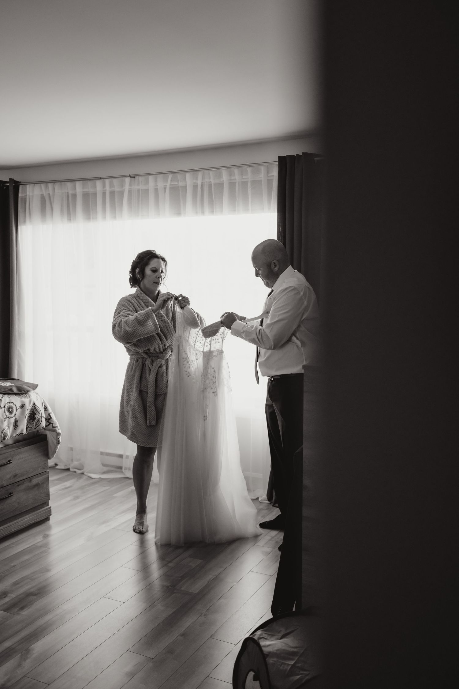 Black and white photo of a bride in a robe being helped into her wedding dress before the ceremony.
