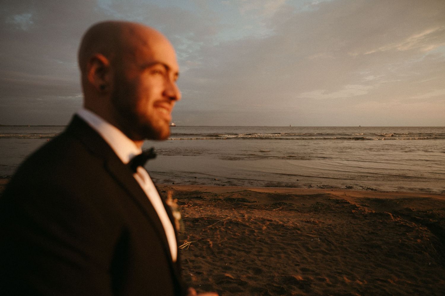 A bald groom in a tuxedo gazes toward the ocean during a golden hour beach wedding.