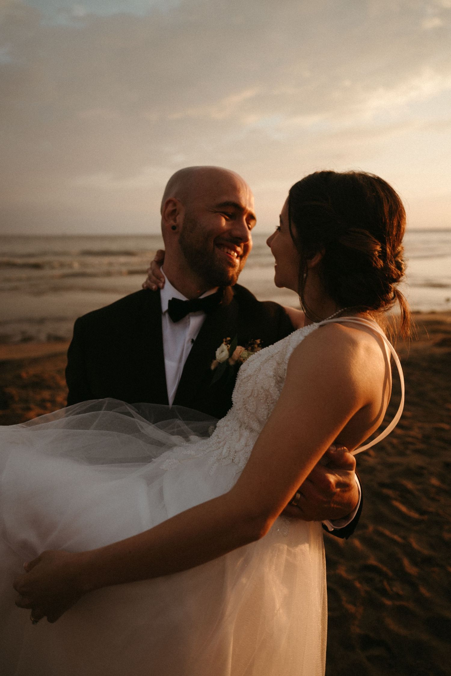 Bride and groom share a romantic embrace on a sandy beach at golden sunset, groom smiling in tuxedo.