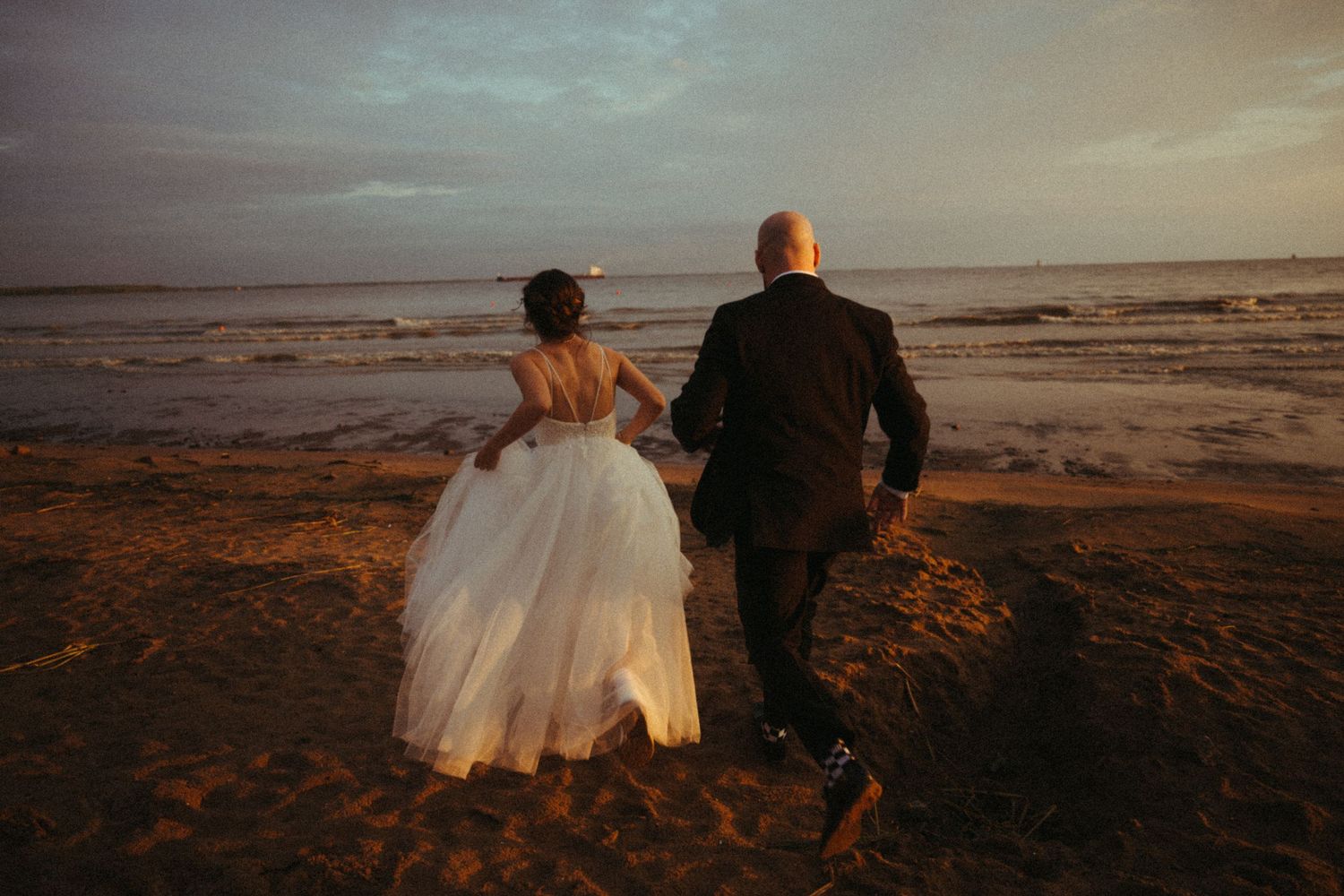 Bride in white gown and groom in black tux running hand-in-hand toward ocean waves at dusk.