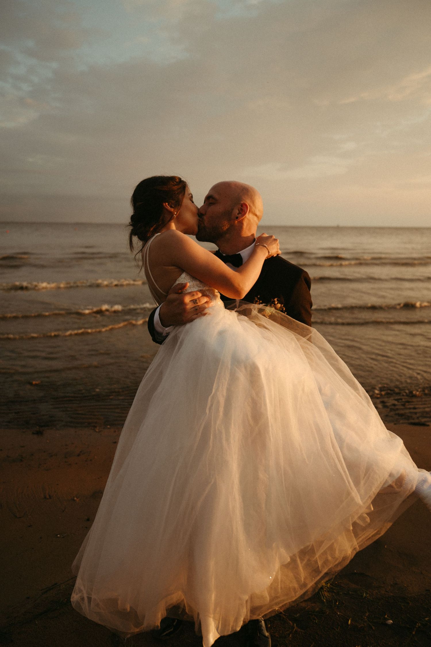 Bride and groom sharing a romantic kiss on the beach at sunset, with ocean waves in the background.