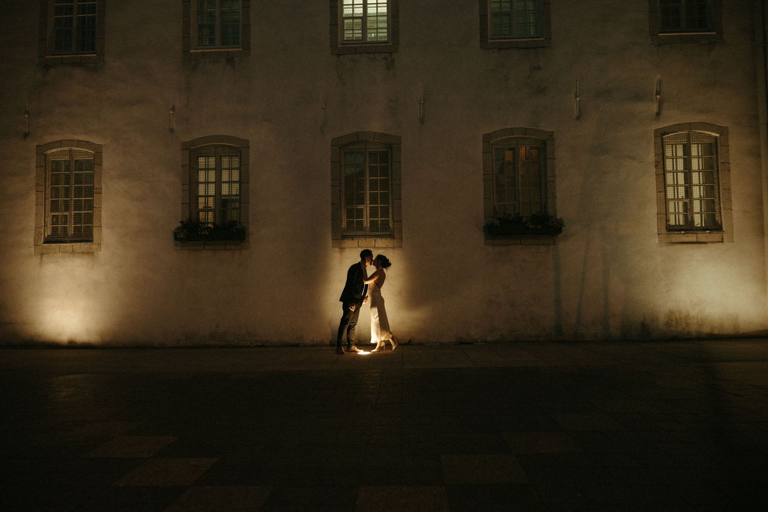 Romantic silhouette of a couple kissing at night against a warmly lit historic stone building facade.