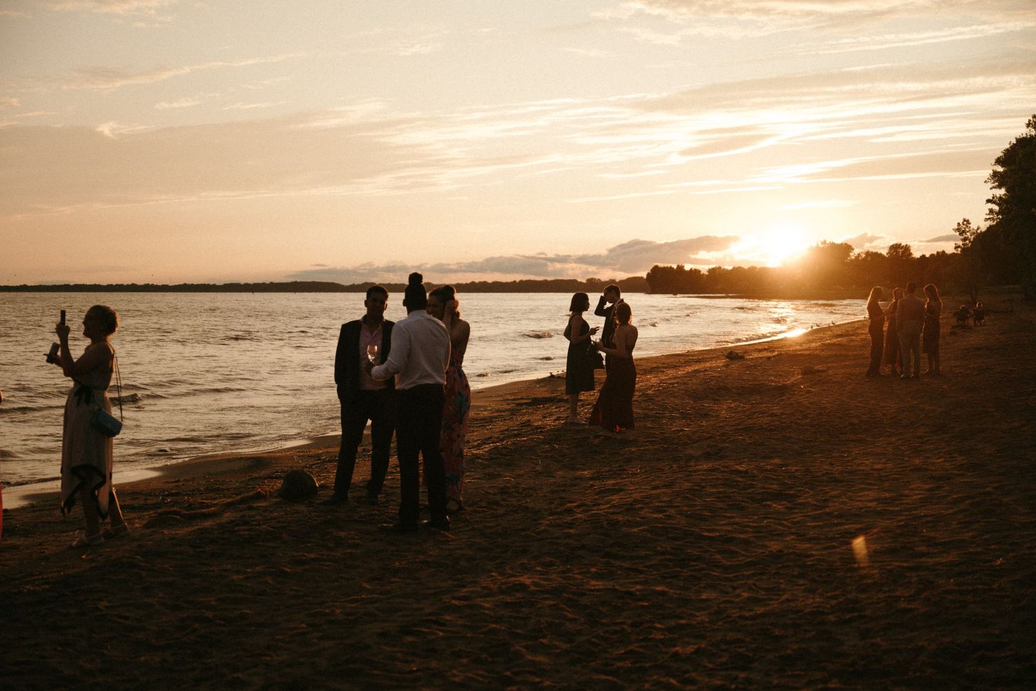Wedding guests gather on a sandy beach silhouetted against a stunning golden sunset.