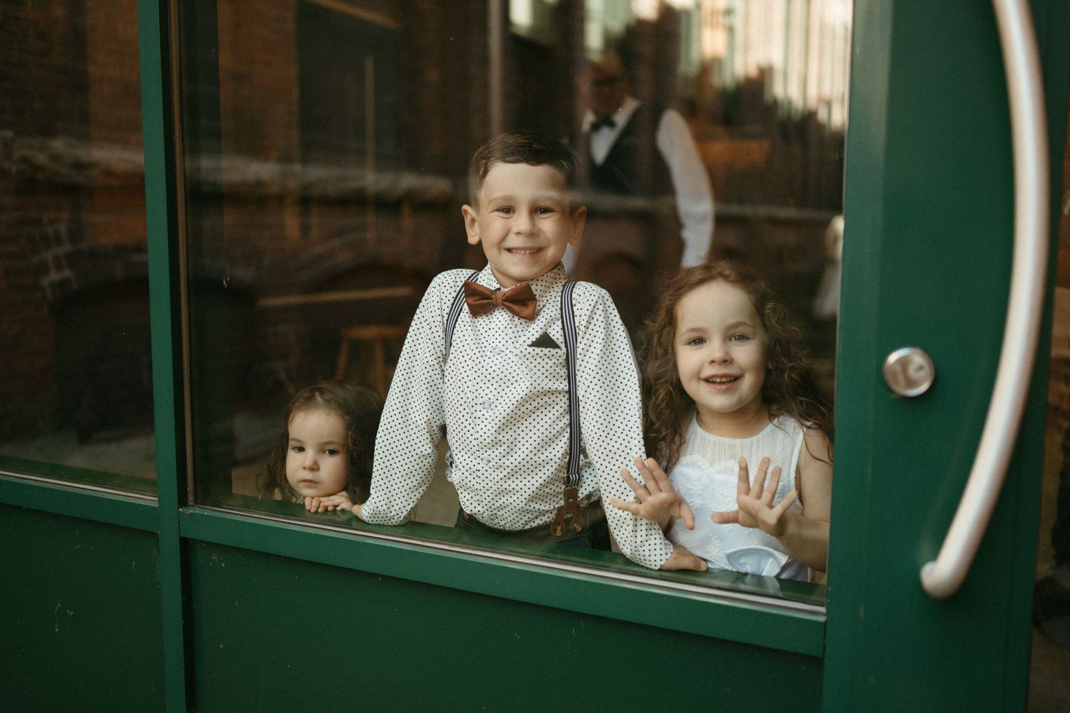 Three children peek through a green-framed glass door, dressed in formal wedding attire.