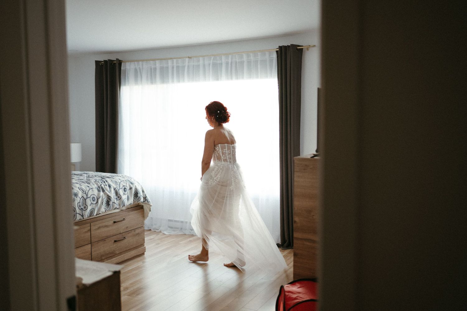 Bride in a flowing white wedding dress standing barefoot in a sunlit bedroom, viewed from a doorway.