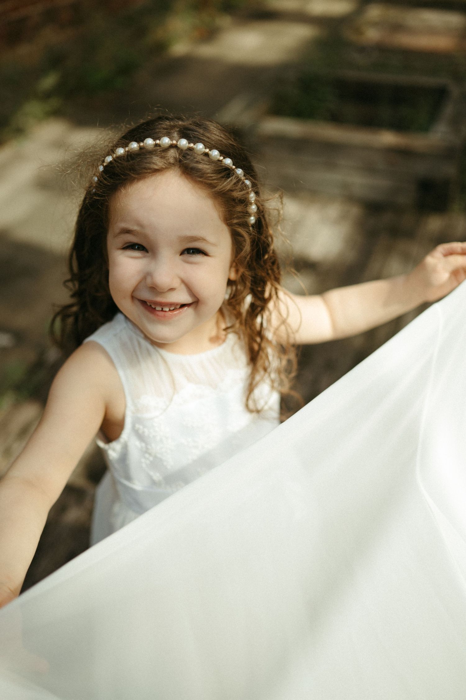 A smiling little girl in a white dress and pearl headband holds a flowing white fabric outdoors.