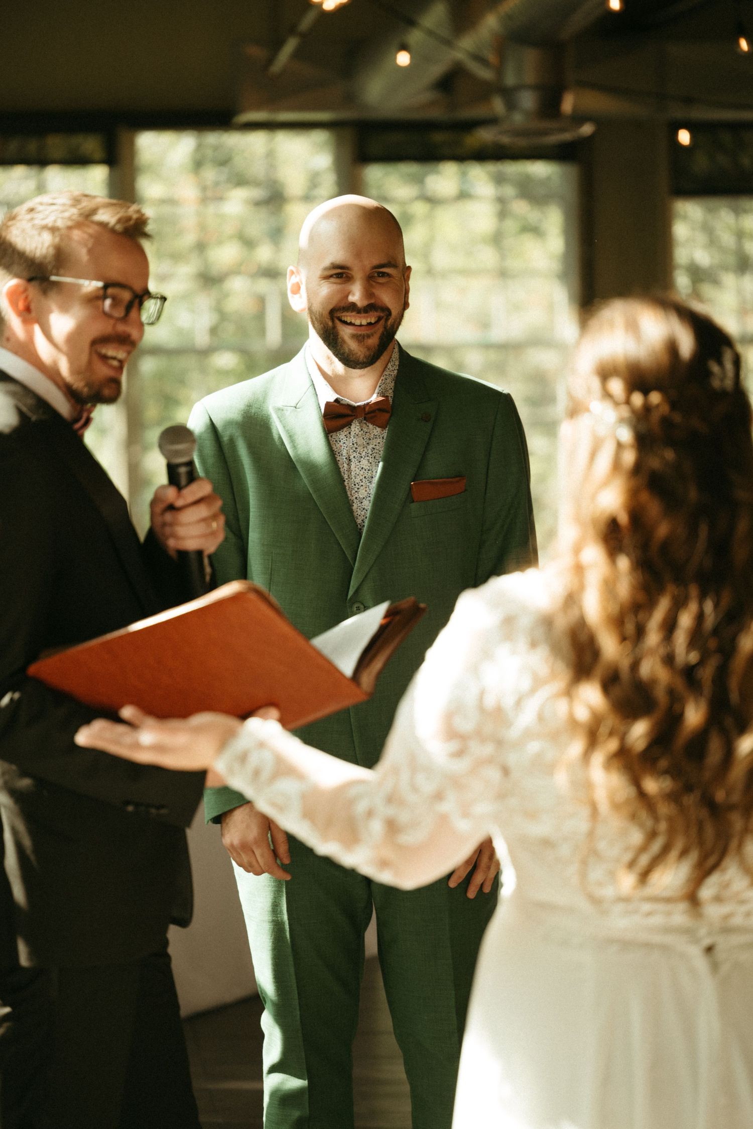 Groom in green suit laughs joyfully during wedding ceremony while officiant holds microphone and bride reads vows.