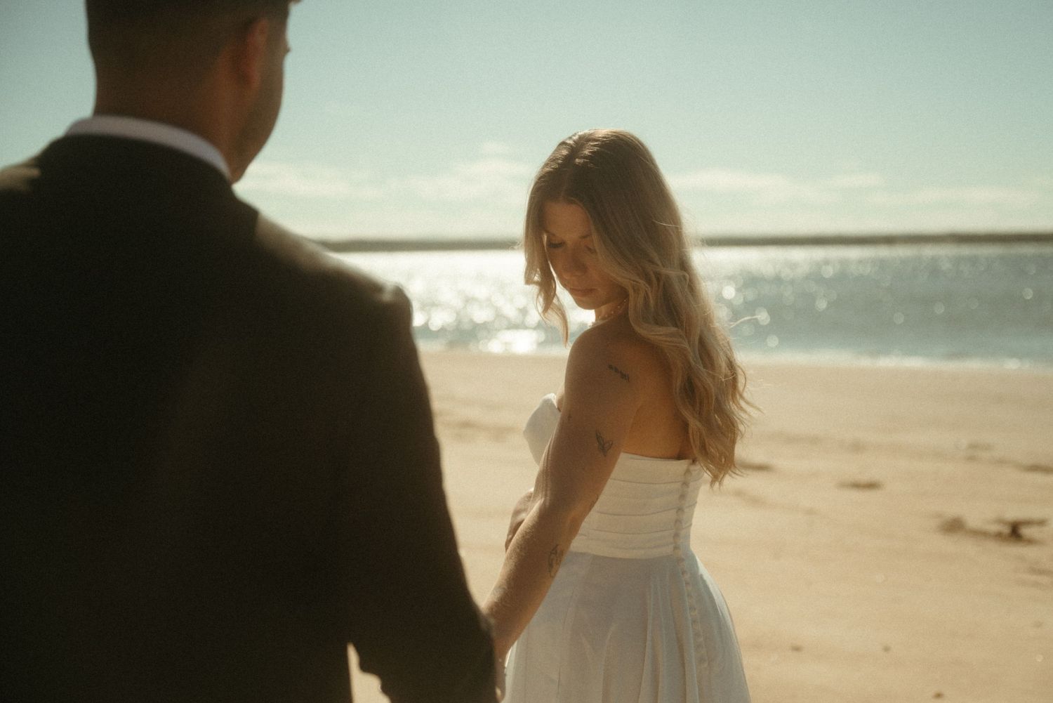 A couple walks hand-in-hand on a sandy beach with the ocean in the background.