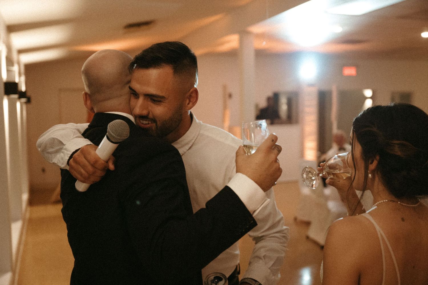 Groom holding a baby while embracing a guest during an indoor wedding reception.