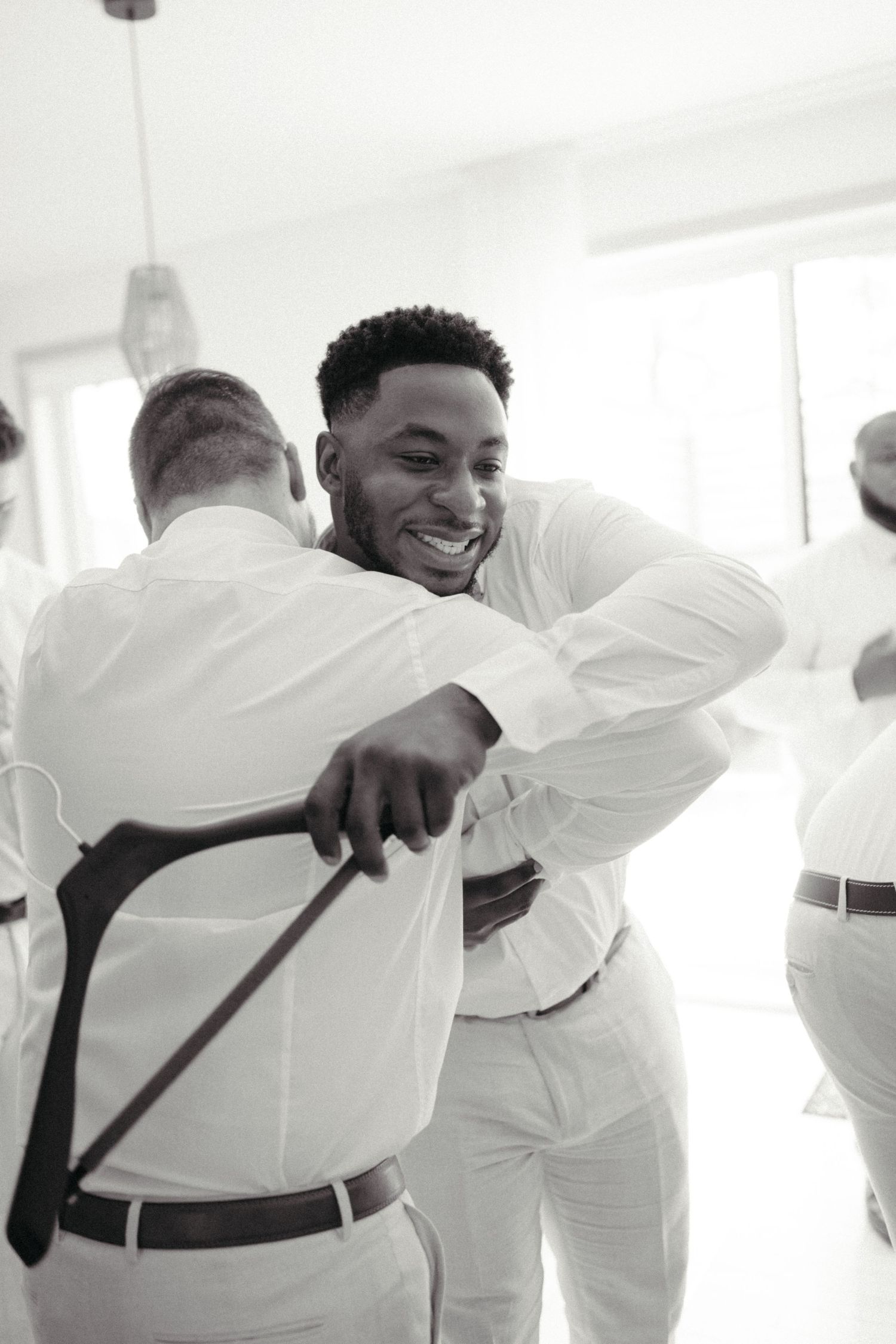 Groom smiling while hugging groomsman in white shirts during wedding preparations, black and white photo.