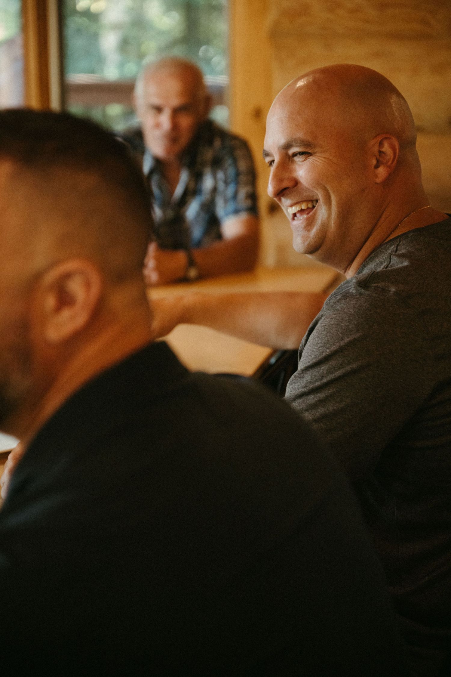 Bald man smiling and laughing during a casual gathering at a wooden cabin table with friends.