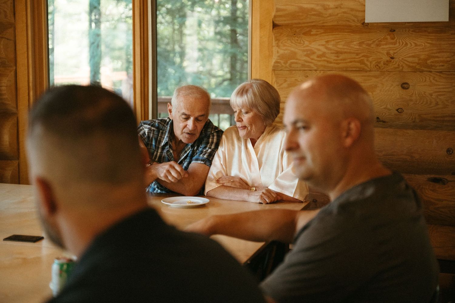 Older couple reviewing documents with two men at a rustic wooden cabin table near a window.