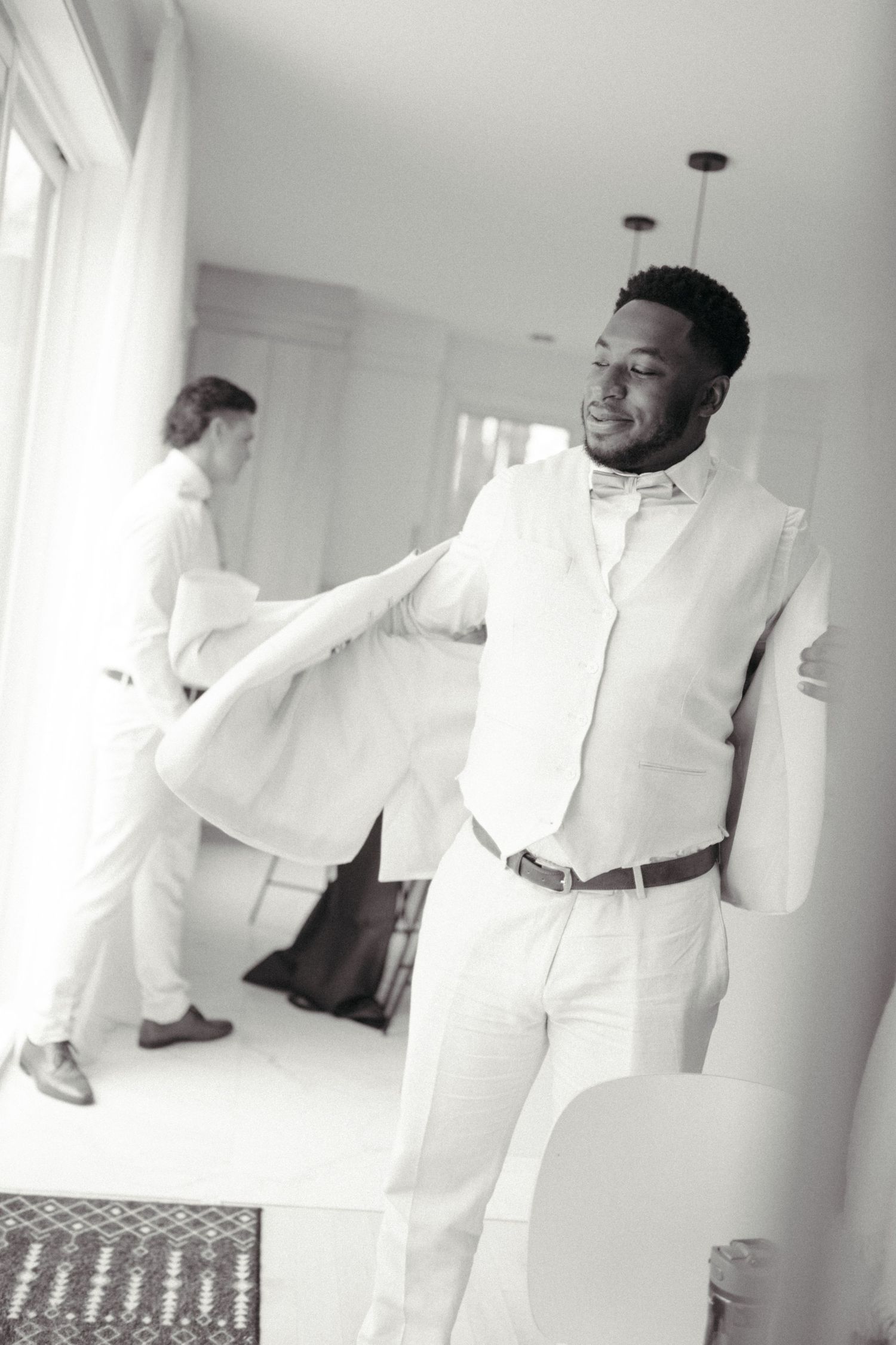 Groom in white suit smiling while getting dressed on wedding day, black and white photo.