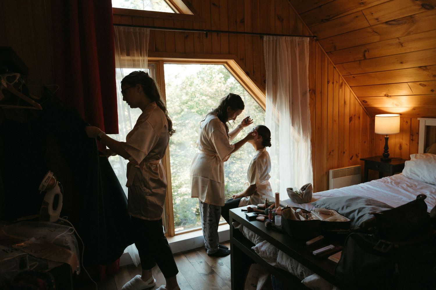 Women in white robes getting ready in a cozy wood-paneled cabin room with natural window light.