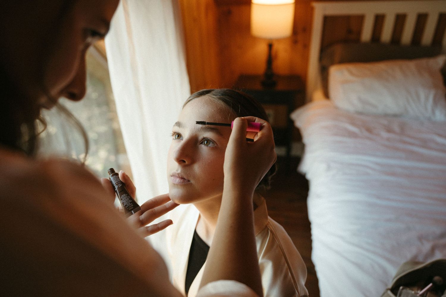 A makeup artist carefully applies eyebrow makeup to a bride getting ready on her wedding day in a cozy bedroom.