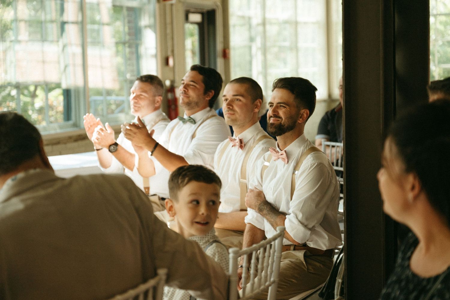 Groomsmen in white shirts with suspenders laugh and clap during a wedding reception at an elegant venue.