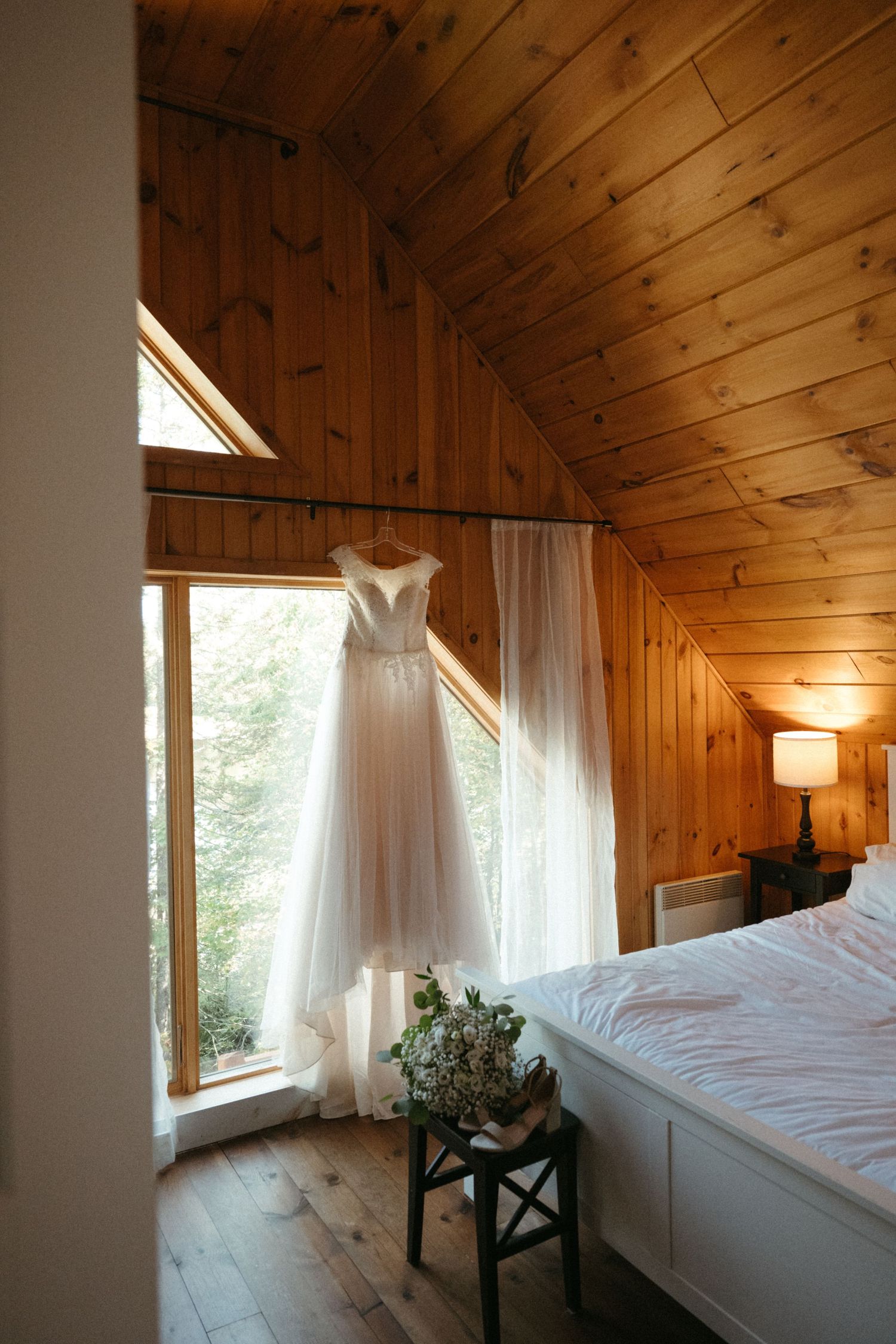 White wedding gown hanging in a rustic wooden cabin bedroom with natural window light.